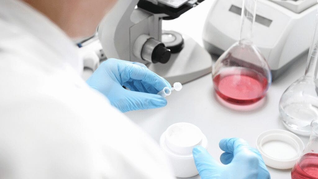 A scientist wearing blue gloves is preparing a sample from a white cosmetic cream jar in a laboratory setting. The workspace includes a microscope, laboratory glassware containing red and clear liquids, and other scientific instruments, indicating cosmetic product research or formulation testing.