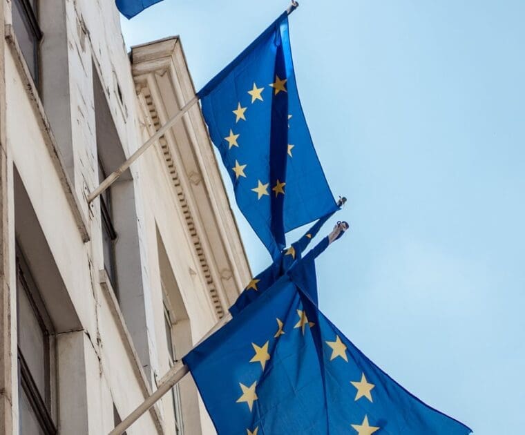 European Union flags with yellow stars on a blue background hanging from a building facade against a clear sky.