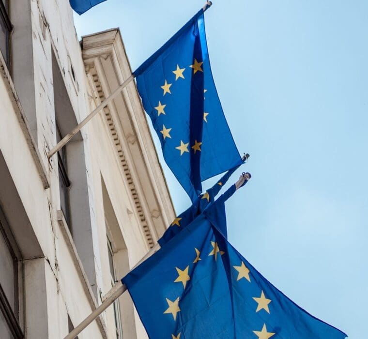 European Union flags with yellow stars on a blue background hanging from a building facade against a clear sky.