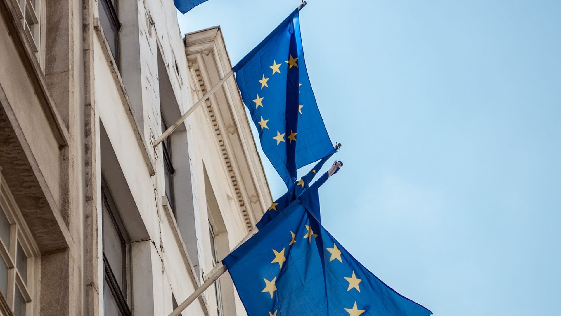 European Union flags with yellow stars on a blue background hanging from a building facade against a clear sky.