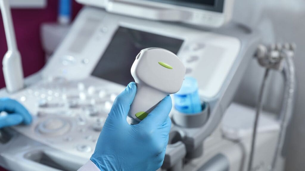 Doctor’s hand in blue glove holding an ultrasound transducer with ultrasound machine in the background.