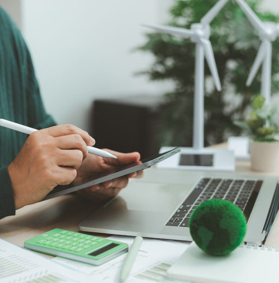 Person using a digital tablet for green energy analysis with wind turbine models and eco-friendly office elements.